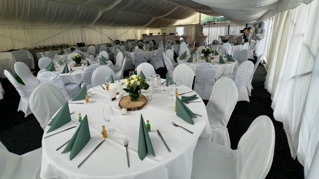 Eleganter Hochzeitstisch im festlich geschmückten Zelt mit weißen Stuhlhussen und grüner Tischdeko