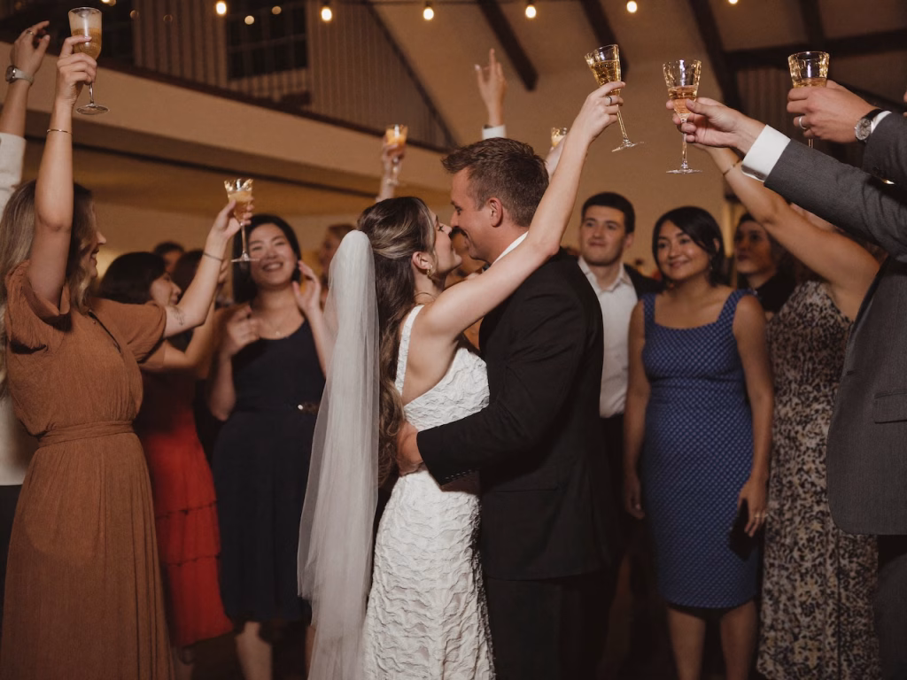 Die Hochzeitsgesellschaft und das Brautpaar stehen auf der Tanzfläche zusammen und stoßen lächelnd mit Sektgläsern an.