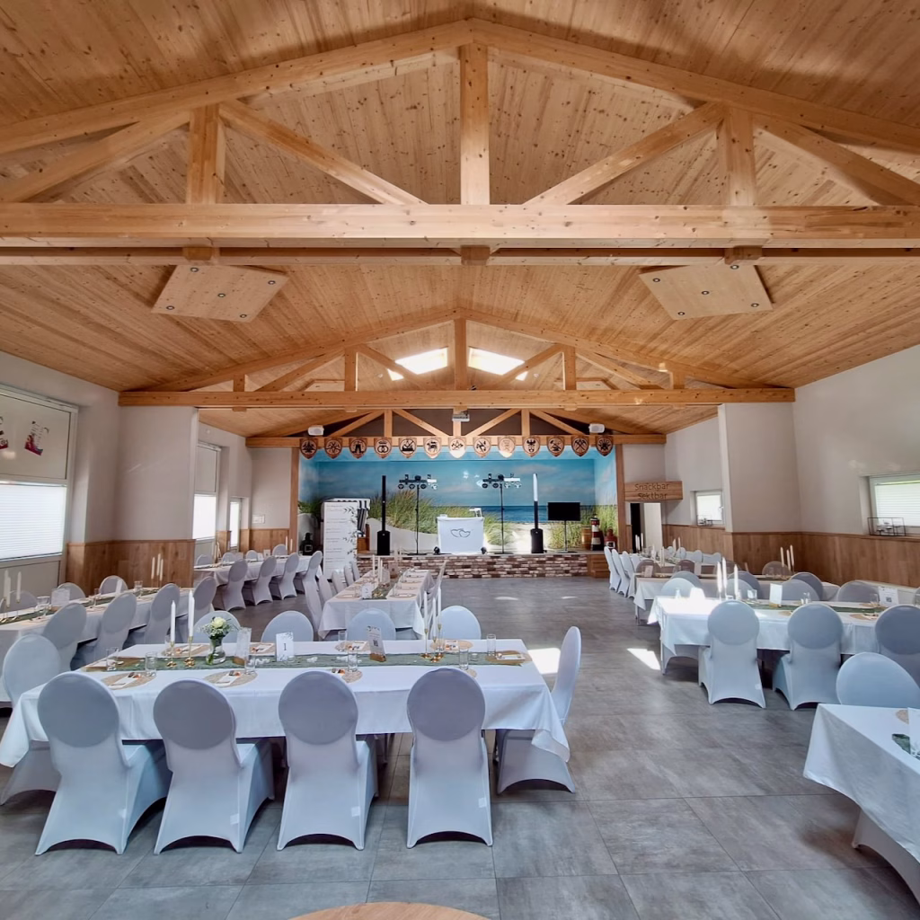 Festlich gedeckte Partyhalle Hooksiel im Wangerland – DJ-Setup auf der Bühne mit Strandbild-Hintergrund, weiß eingedeckte Tische mit Holzdachstuhl