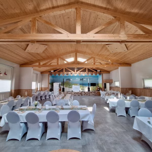 Festlich gedeckte Partyhalle Hooksiel im Wangerland – DJ-Setup auf der Bühne mit Strandbild-Hintergrund, weiß eingedeckte Tische mit Holzdachstuhl