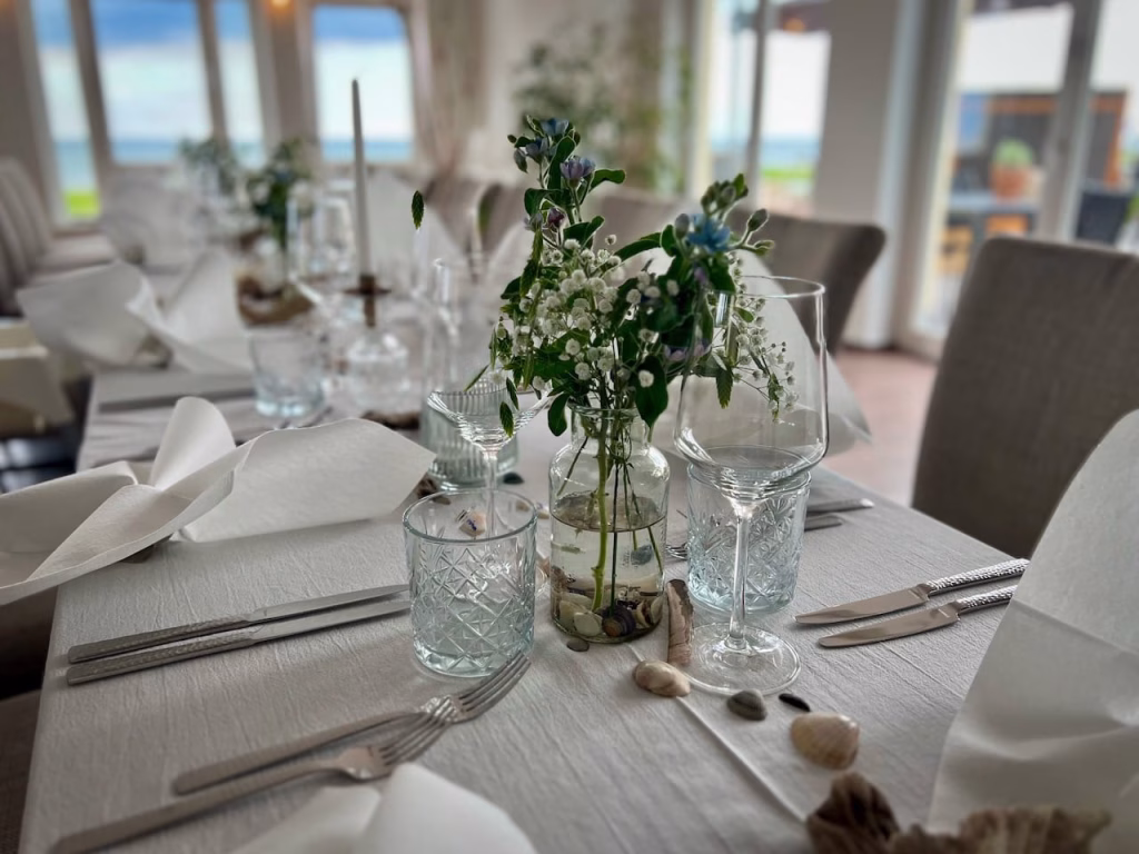 Festlich gedeckter Tisch im Restaurant Krüsch auf Wangerooge – Wildblumen in Glasflasche, Schliffgläser, Muscheln und Meerblick im Hintergrund