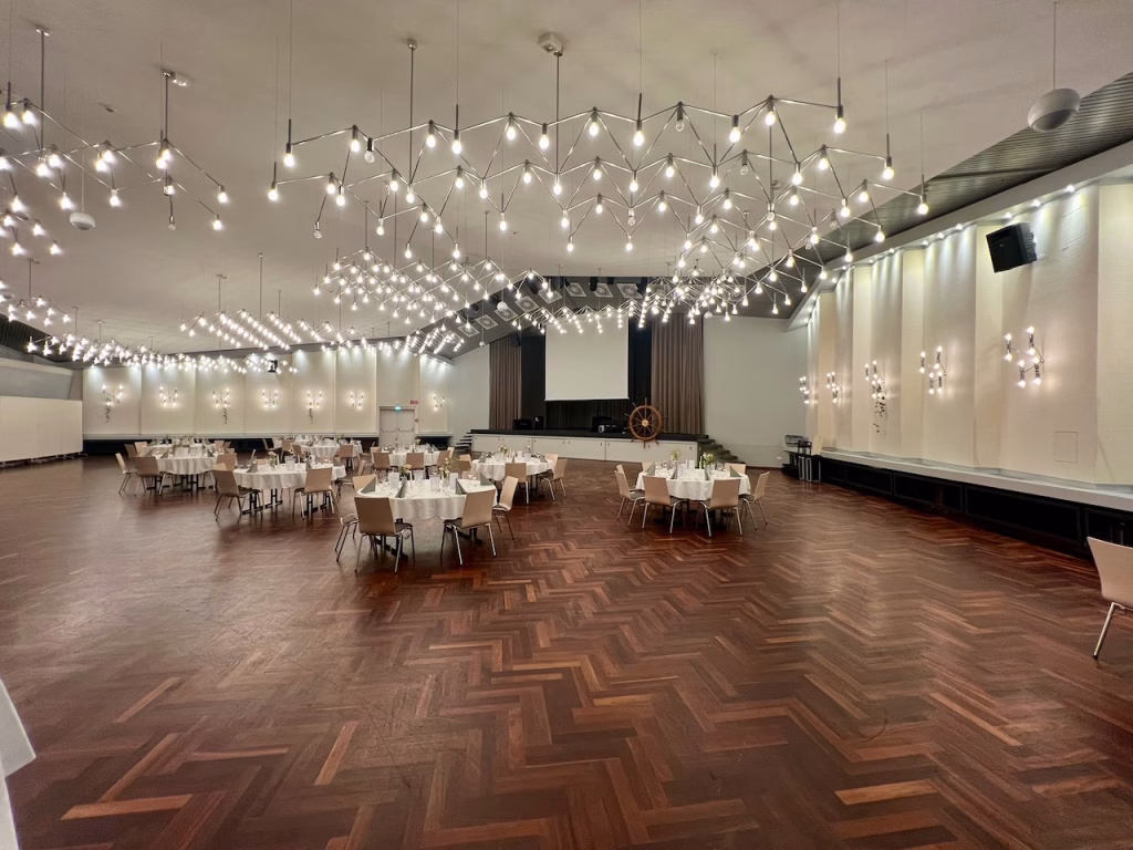 Großer Saal im Gorch-Fock-Haus Wilhelmshaven mit Lichtdesign, Parkettboden und locker verteilten runden Tischen