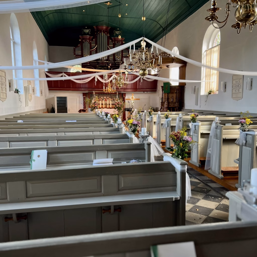 Festlich geschmückter Innenraum der St. Stephani- und Bartholomäi Kirche in Detern für eine Hochzeit am 05.09.2026 – weiße Stoffbahnen, Blumen an den Kirchenbänken, Gottesdiensthefte auf den Bänken